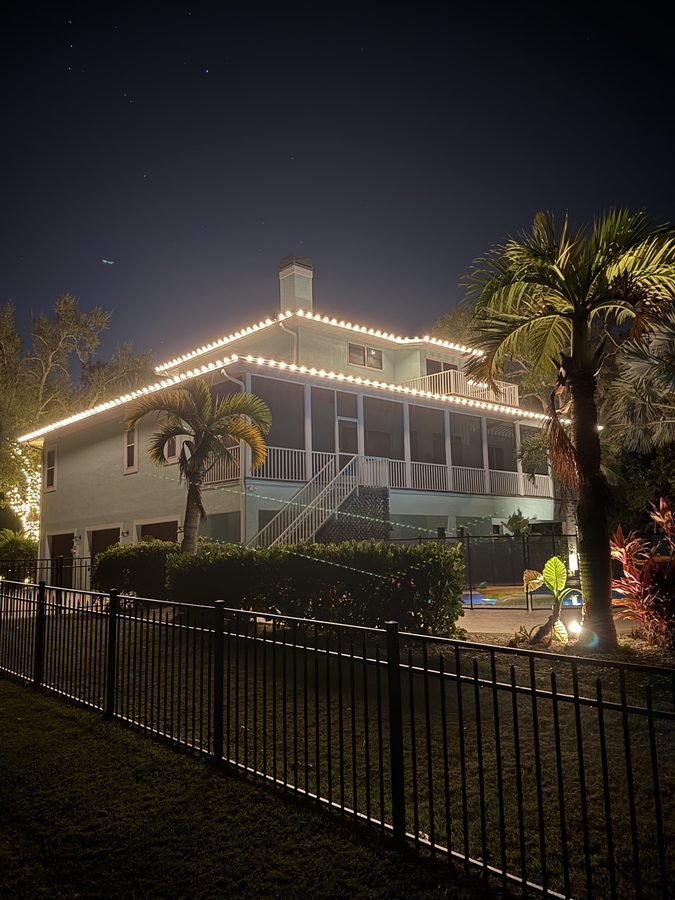 Waterfront ranch-style home in Bradenton with warm white roofline lighting and a wrapped palm tree at dusk