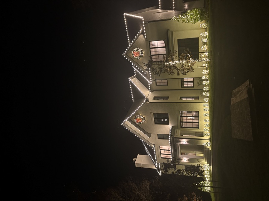 Cream stucco estate with roofline lighting, wreaths, and landscape bed lighting