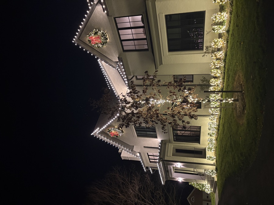Stucco estate with lit wreaths on the gables and a wrapped tree at the entry